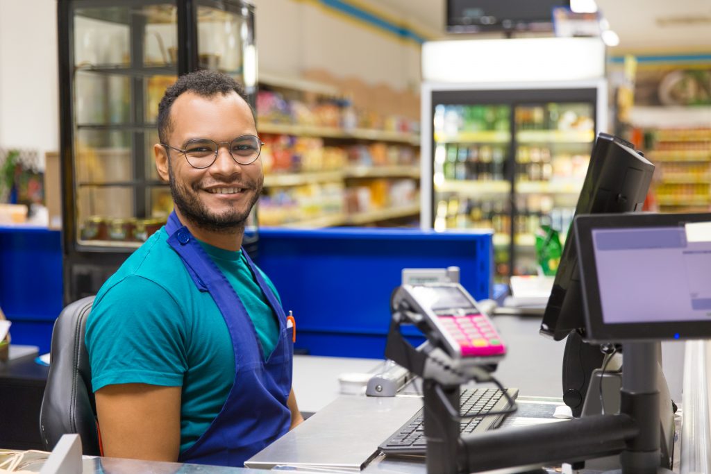 A young man wearing an apron smiles at the camera while seated behind a monitor at a grocery store checkout.